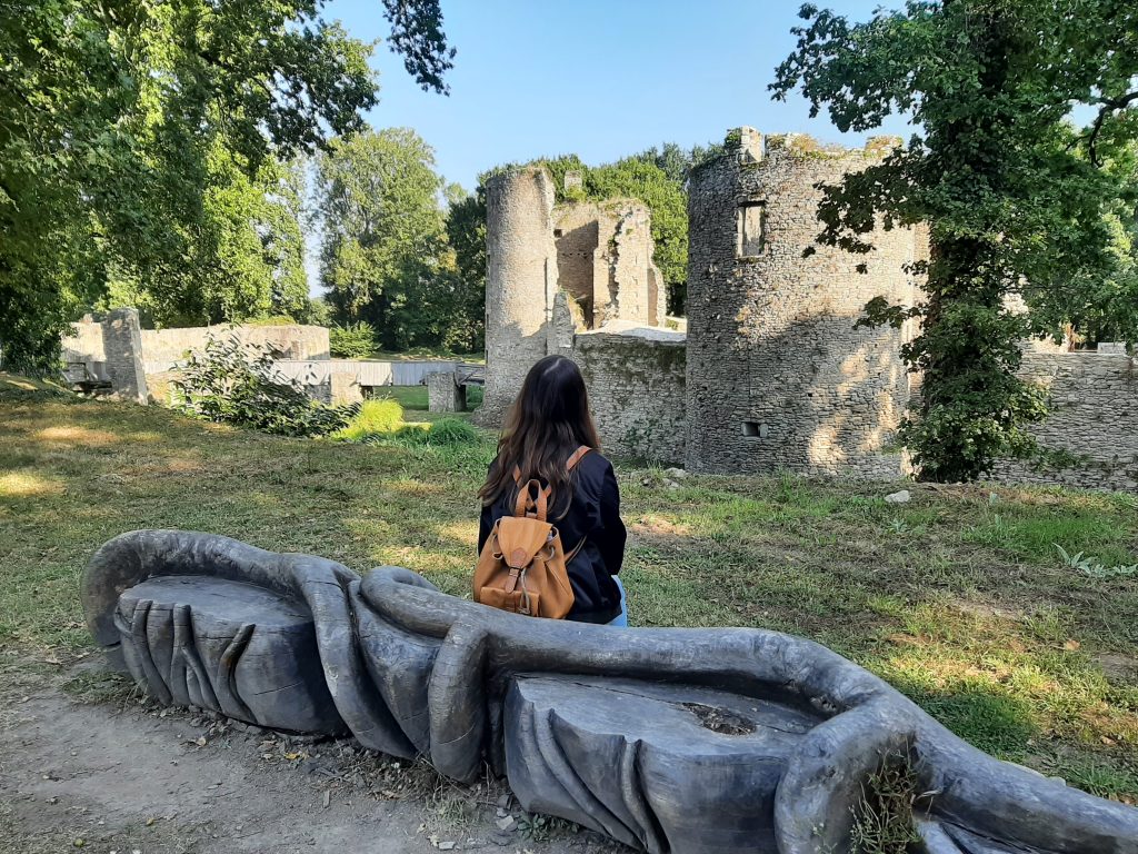 Une femme seule assise sur une sculpture en bois, devant les douves du château.